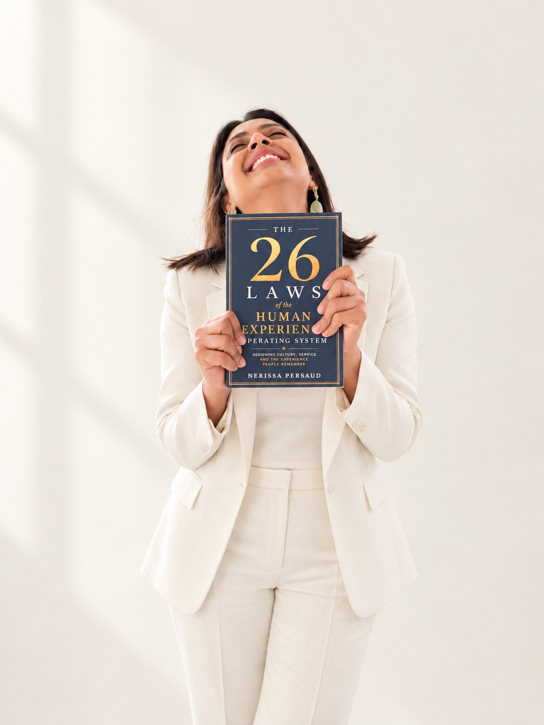 Nerissa Persaud in an ivory suit, joyfully holding The 26 Laws against a soft ivory studio background